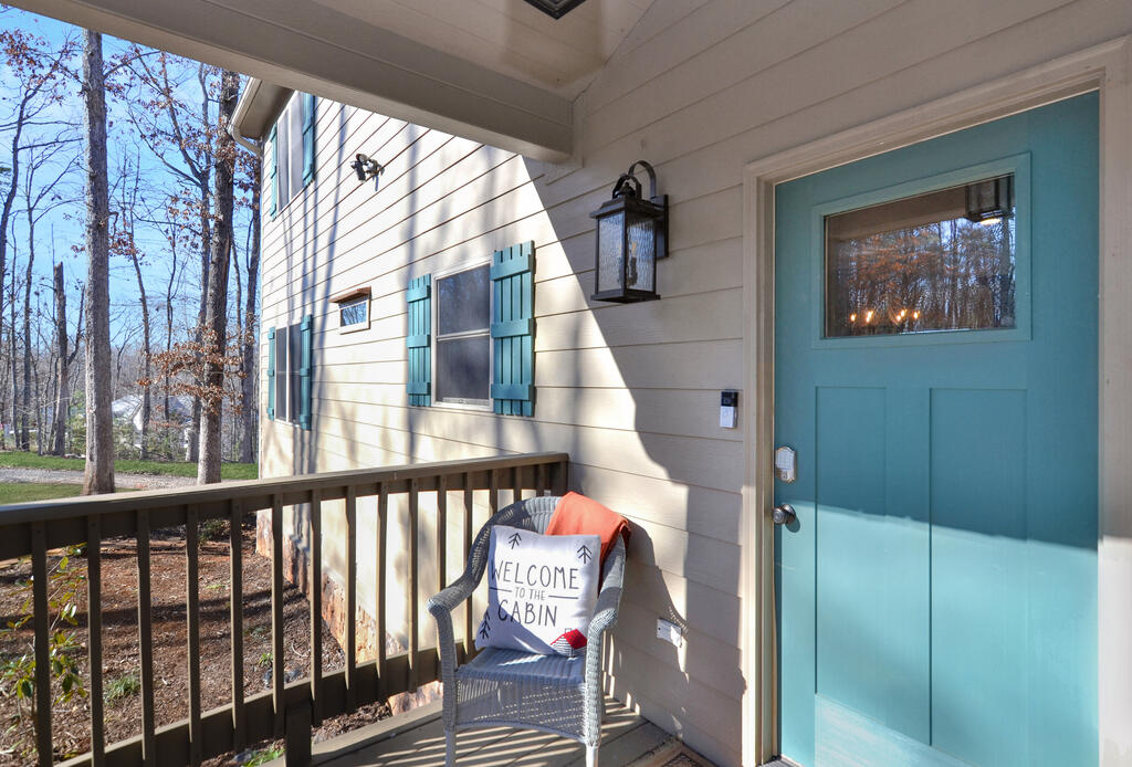 Cabin porch with a turquoise front door and a wicker chair holding a welcome pillow.