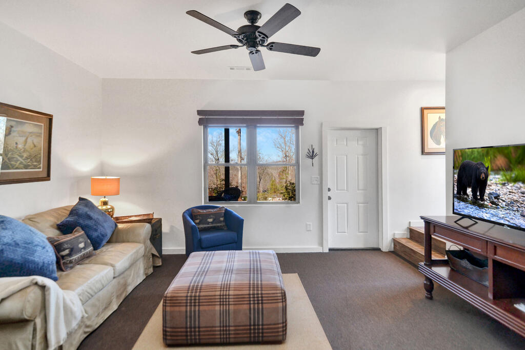 Furnished living room with a plaid ottoman, ceiling fan, and window overlooking trees.