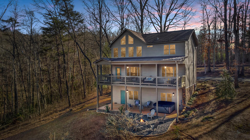 Multi-level gray house among leafless trees, with covered decks, a hot tub, and a fire pit at dusk.