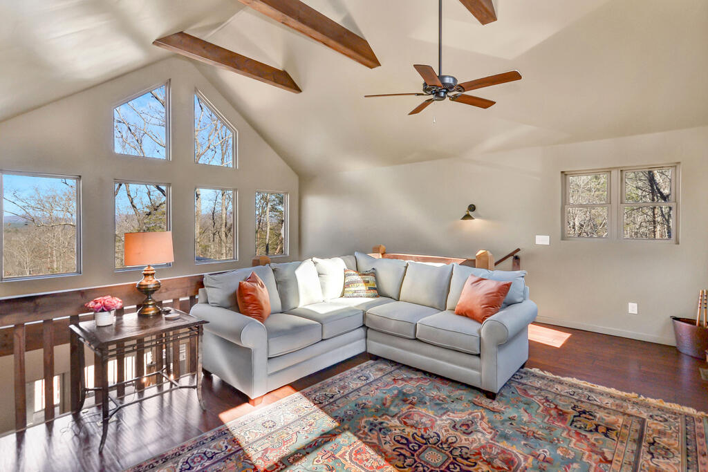 Living room with a gray sectional beneath exposed beams and tall windows overlooking wooded hills.