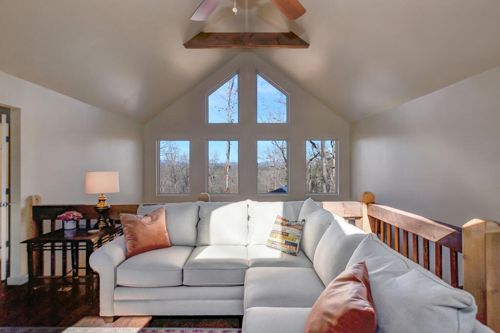Vaulted living room with a gray sectional, wood railing, and tall windows overlooking wooded hills.