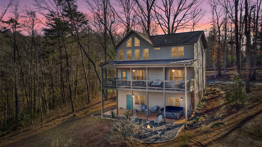 Two-story gray house with covered decks, a hot tub, and fire pit among leafless trees at dusk.