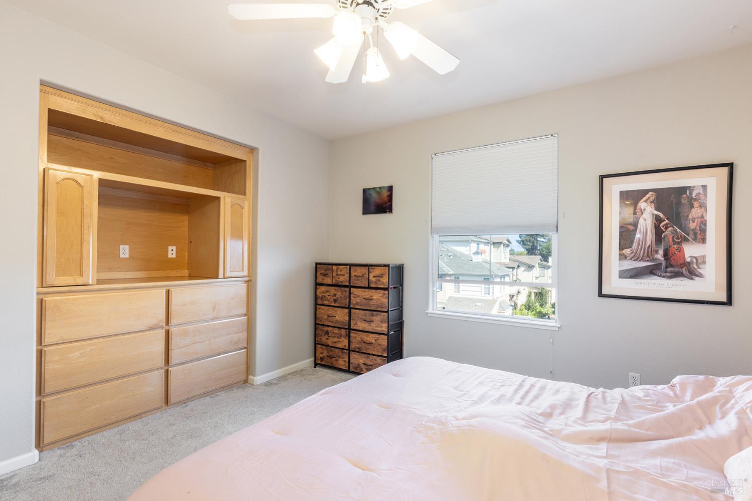 Bedroom with built-in light wood drawers, pale pink bedding, and a window overlooking nearby homes.