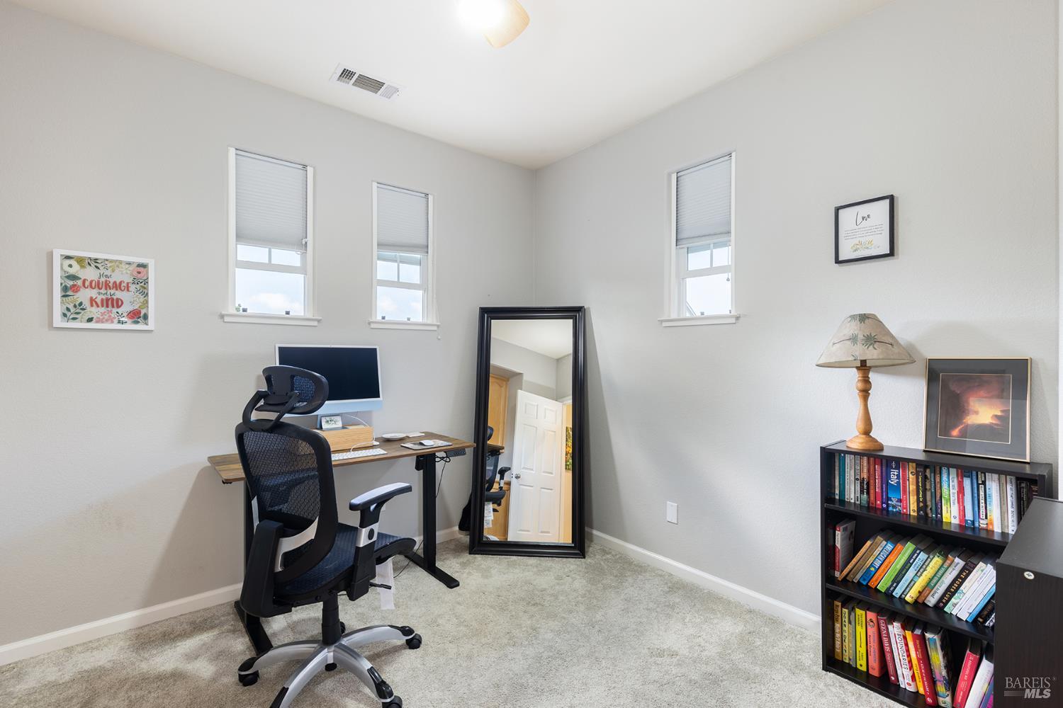 Home office with a desk and chair, full-length mirror, and bookcase beneath high windows.