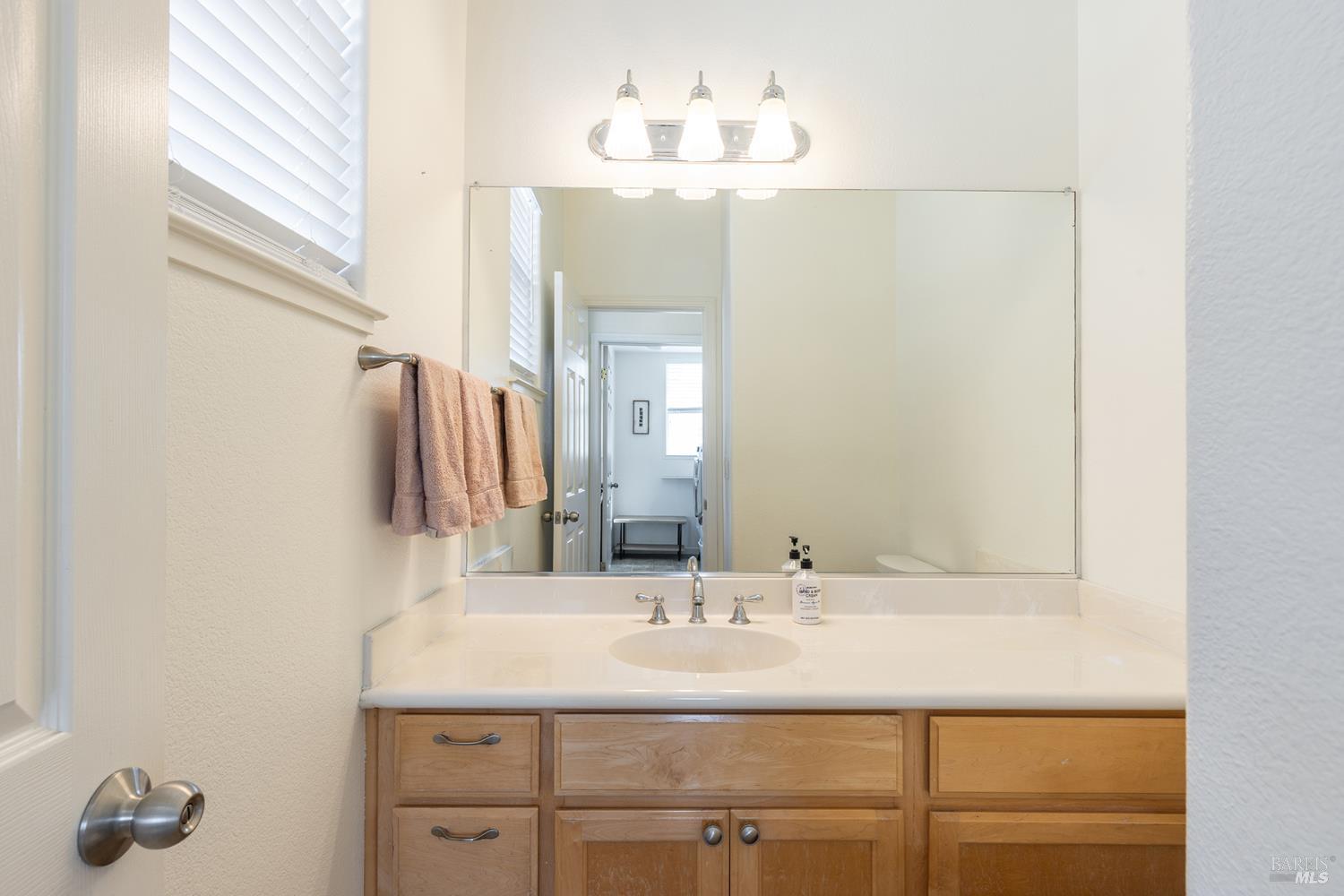 Bathroom vanity with a wide mirror, light wood cabinets, pink towels, and a window with white blinds.