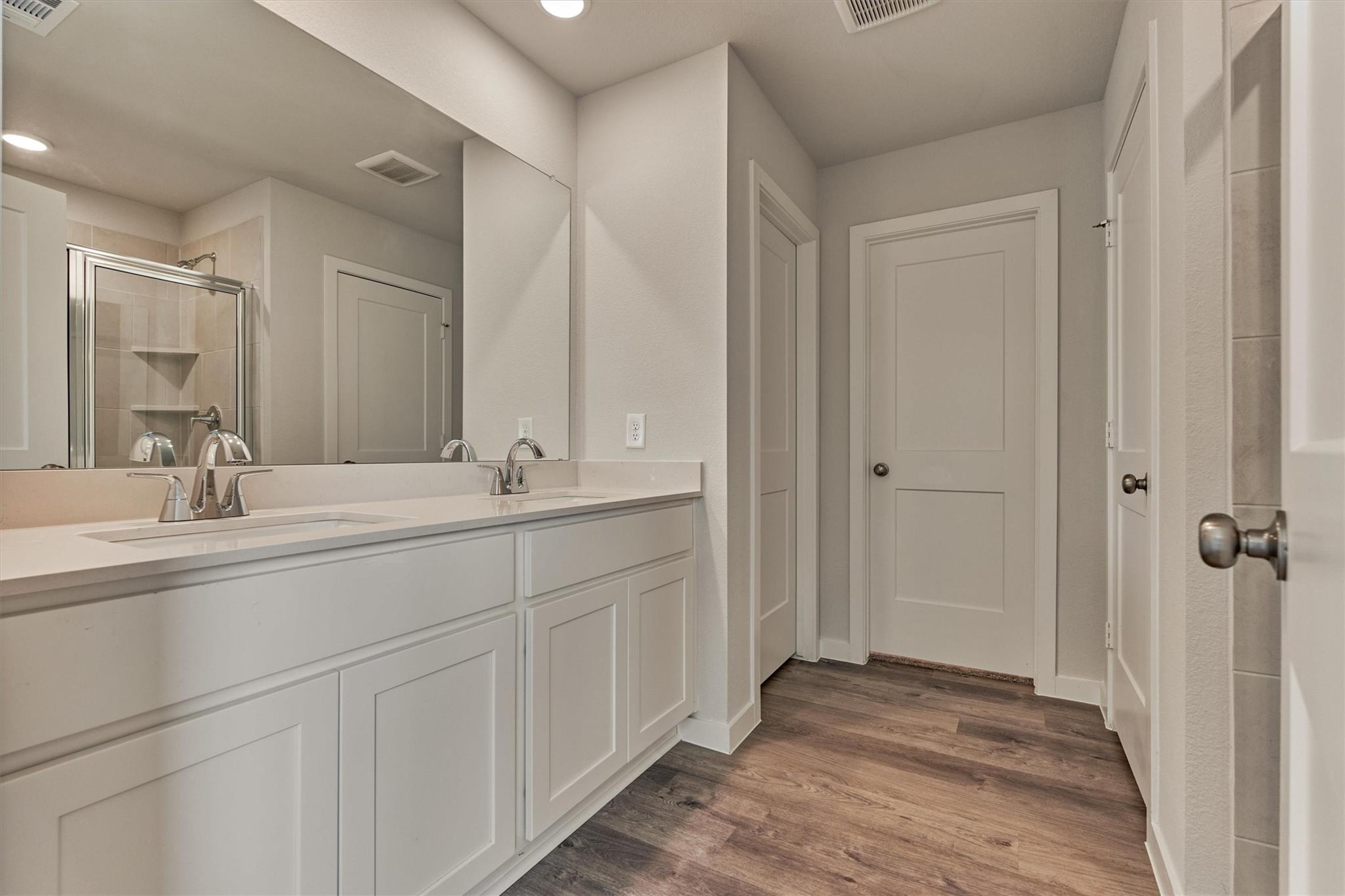Bathroom vanity with a large mirror, glass-enclosed tiled shower, and dark wood-look flooring.