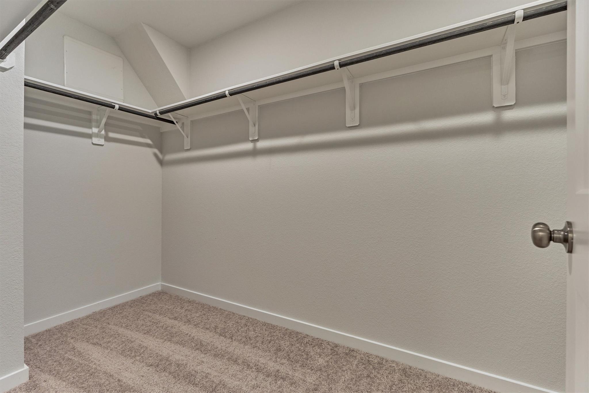 Empty walk-in closet with beige carpet and wraparound clothes rods and shelves.