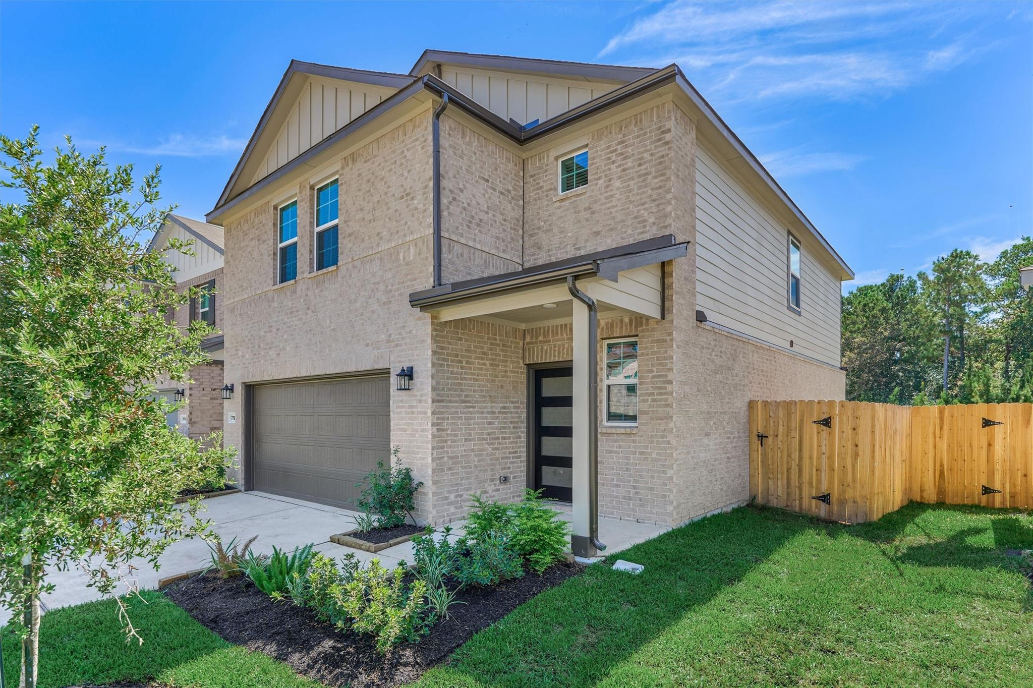 Two-story light-brick house with a covered entry, garage, landscaped lawn, and wooden side gate.