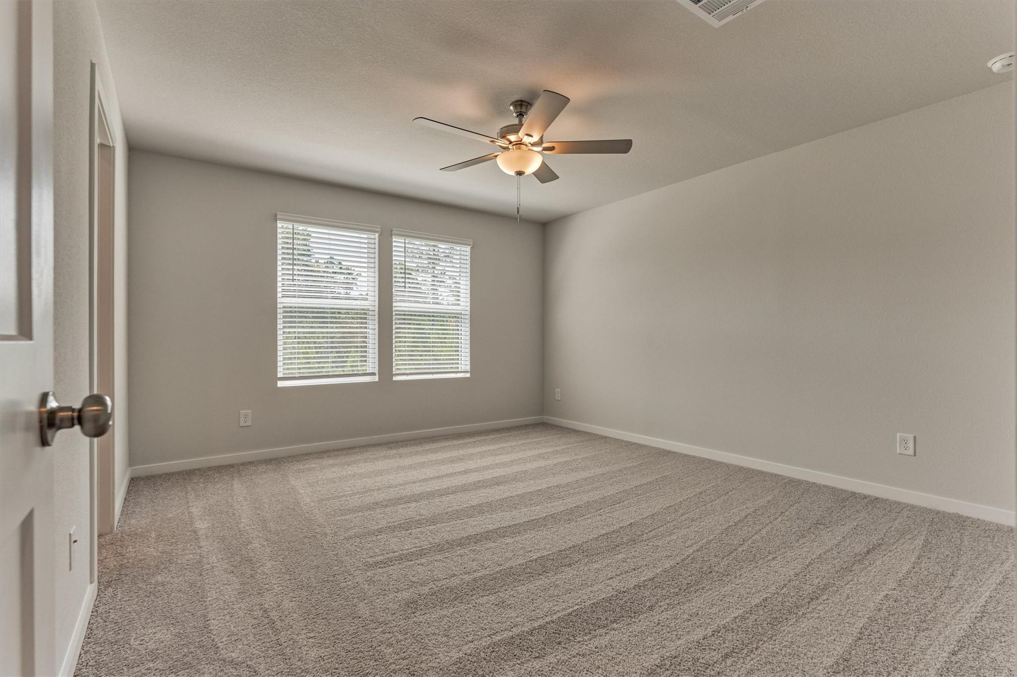 Empty bedroom with beige carpet, two blind-covered windows, and a lit ceiling fan.