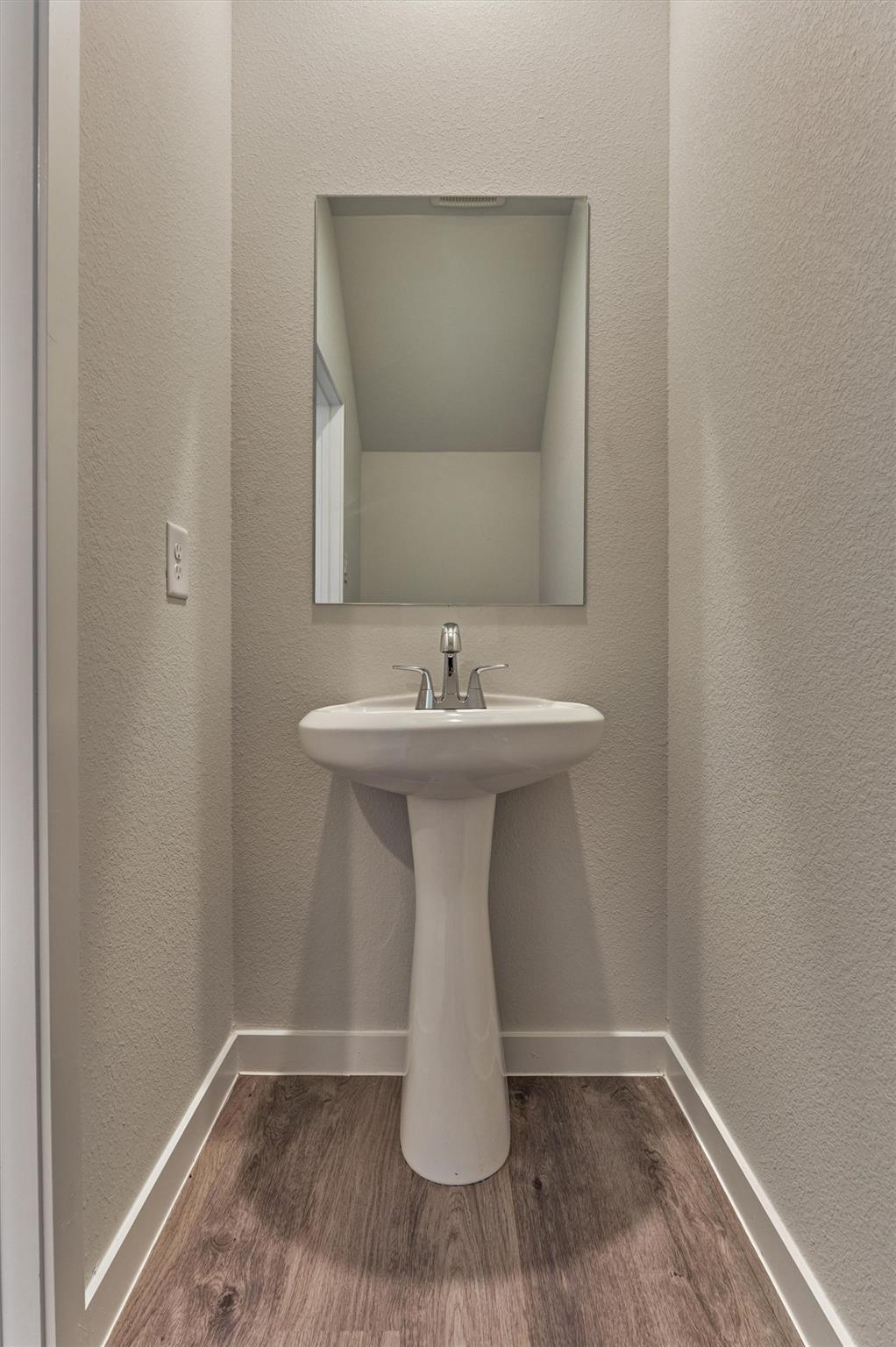 Compact bathroom with a white pedestal sink, rectangular mirror, and wood-look flooring.