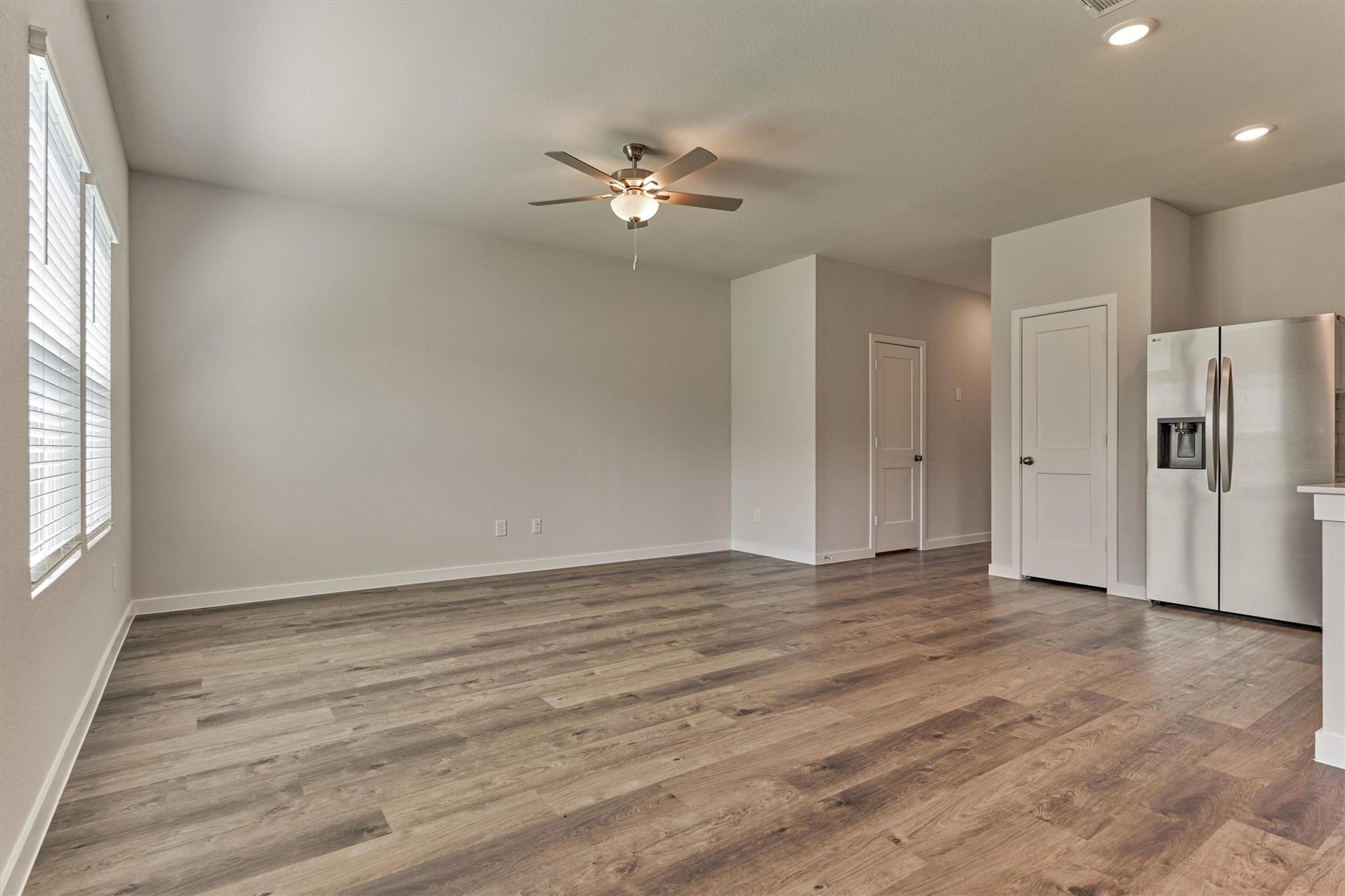Empty open-plan room with wood-look flooring, window blinds, ceiling fan, and stainless steel refrigerator.