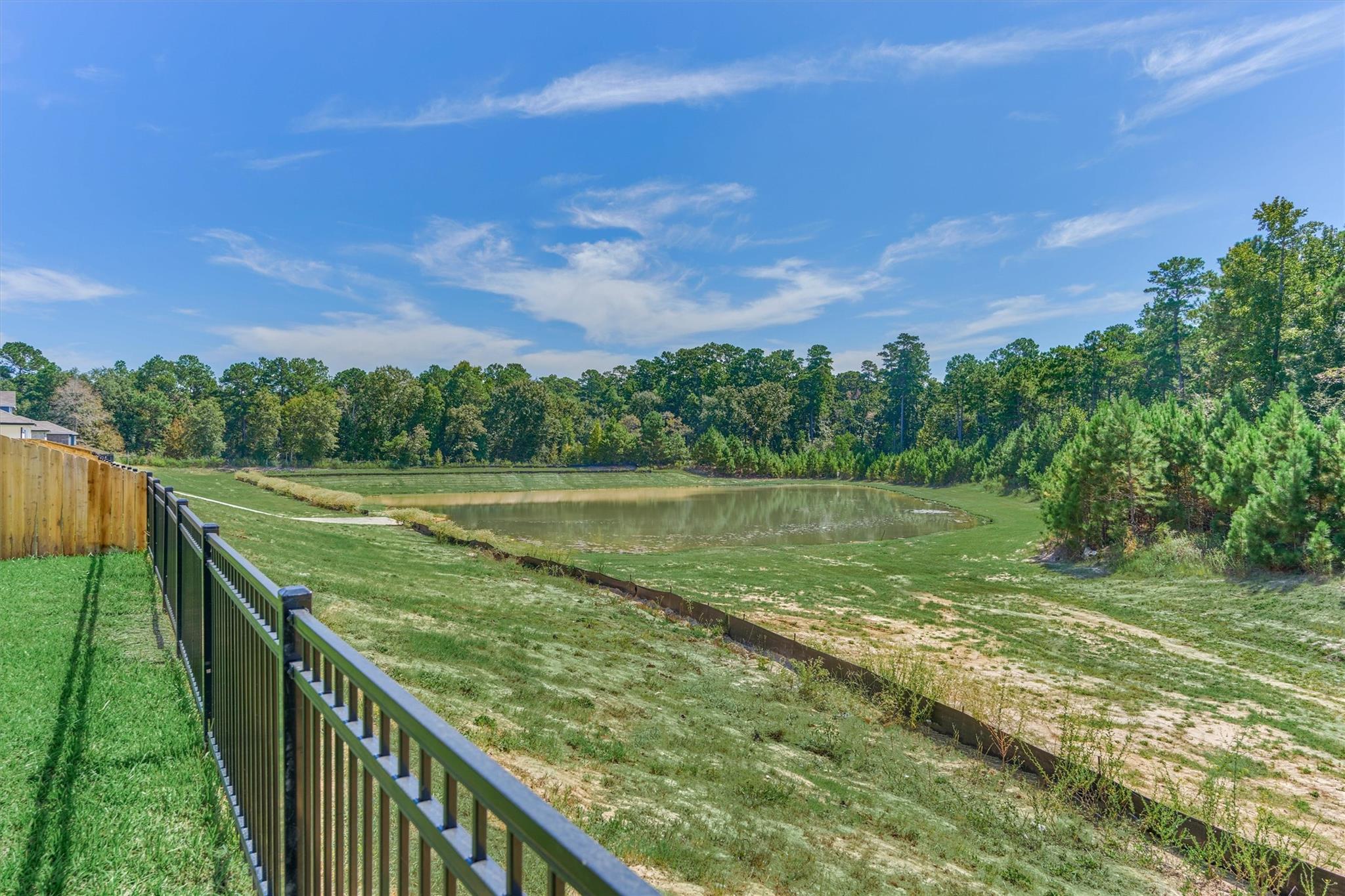Yard overlooking a pond, with a black metal fence and wooded trees beyond.