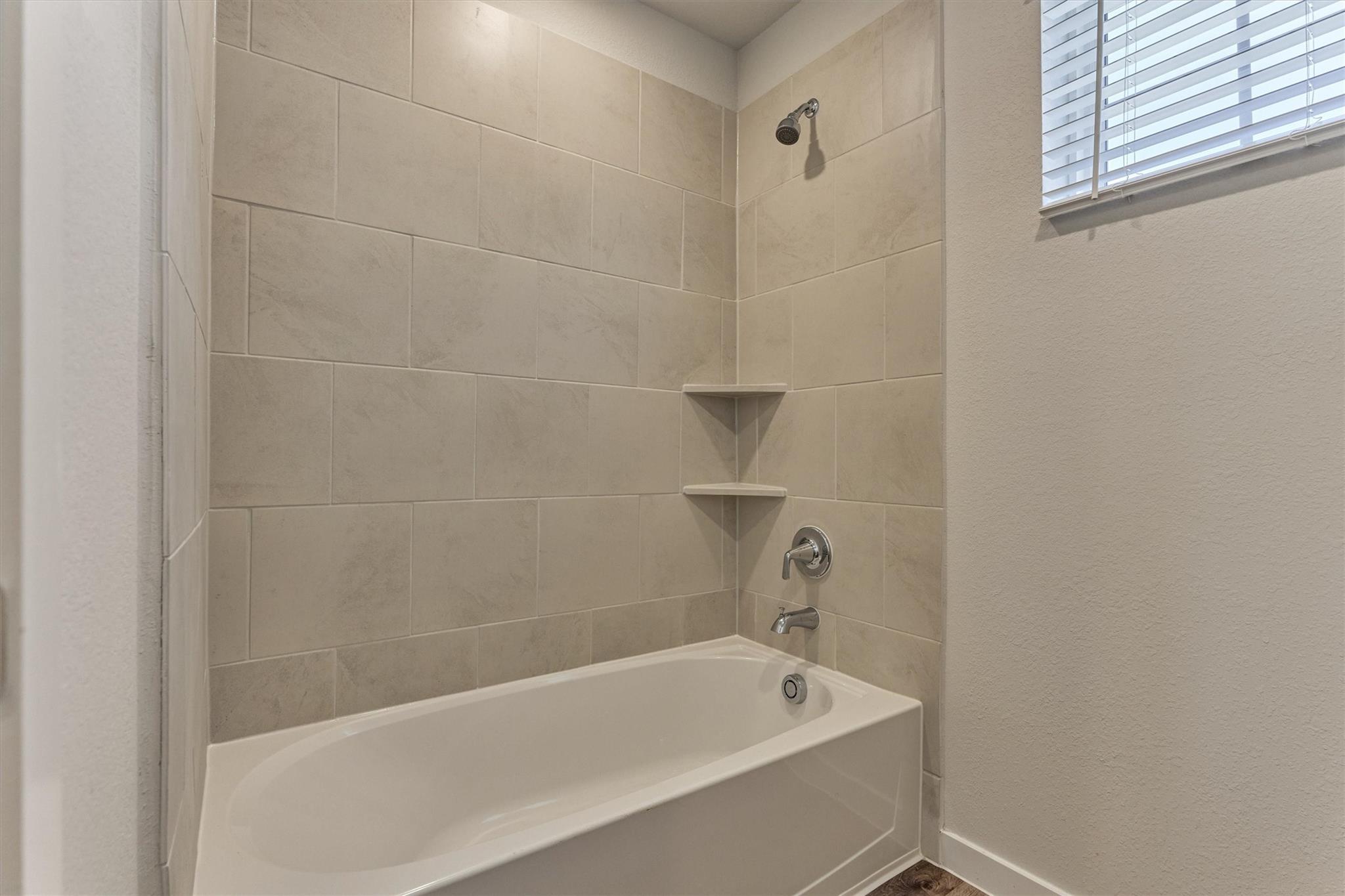 Bathroom with a white bathtub, beige tile surround, and built-in corner shelves.
