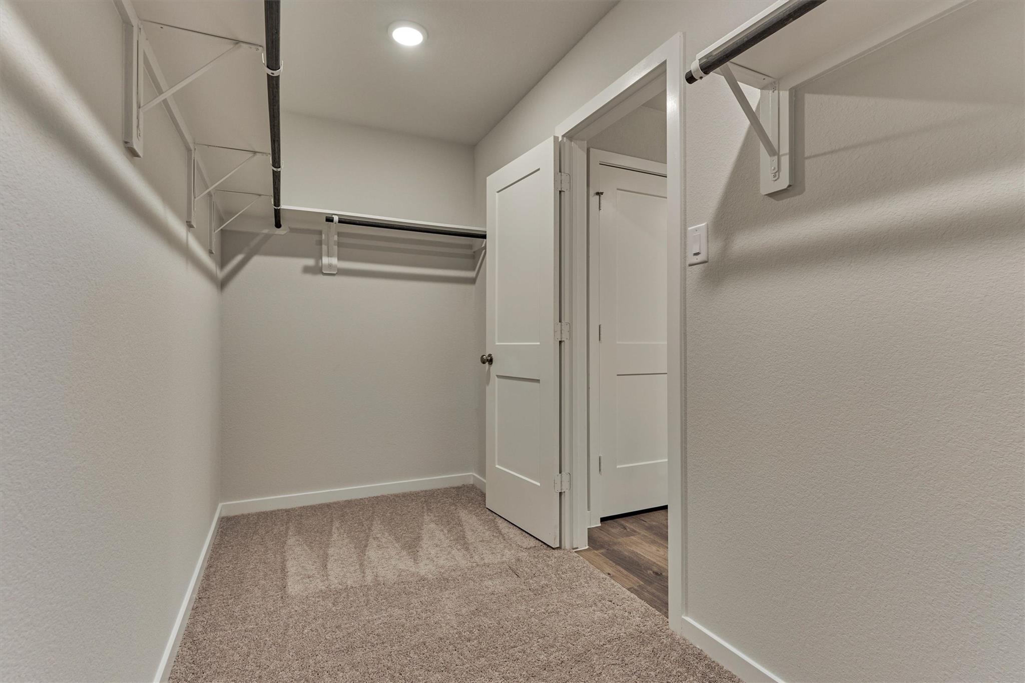 Empty walk-in closet with beige carpet, black hanging rods, and an open door to a wood-floored hallway.