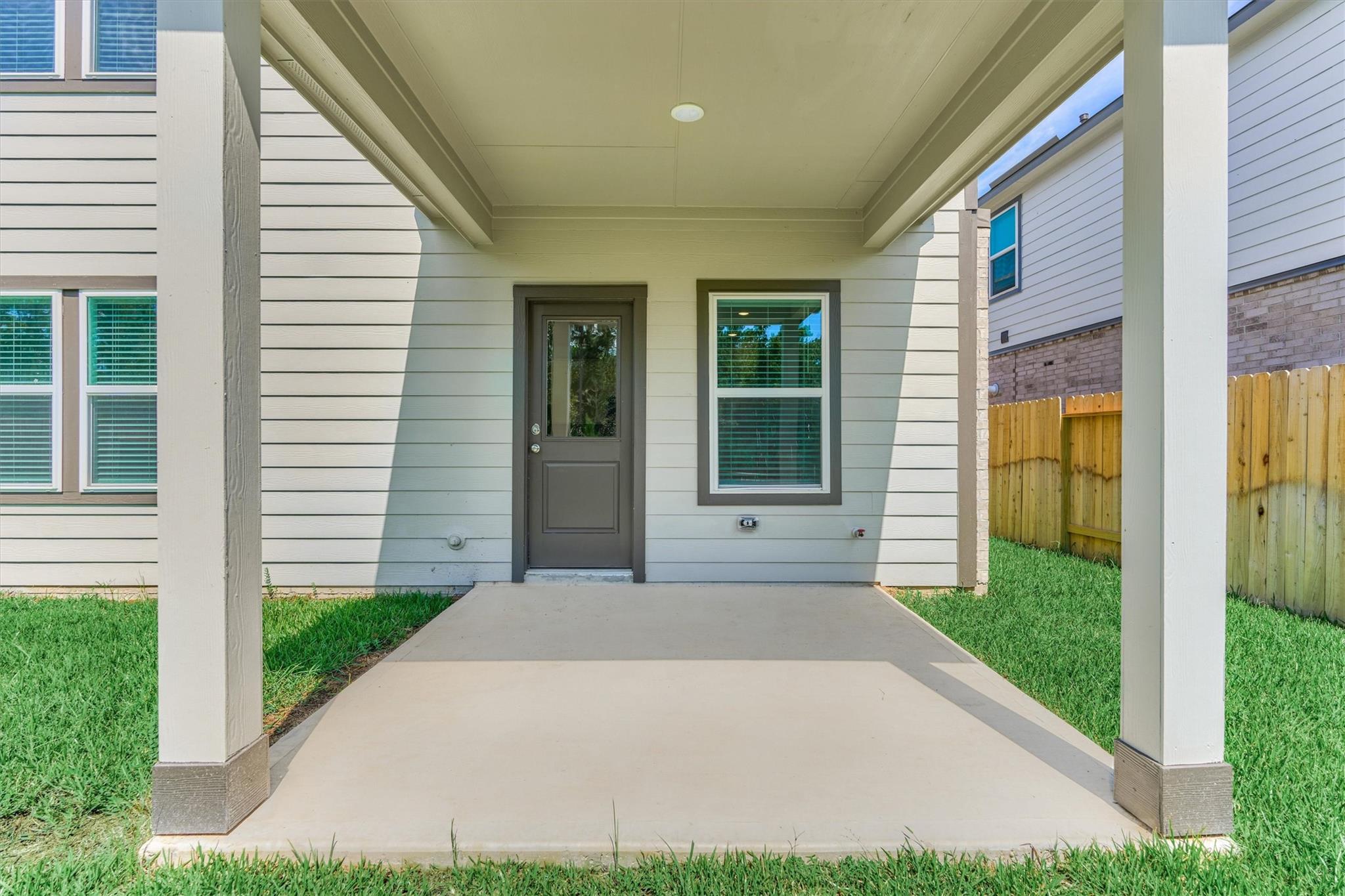 Covered concrete patio with a gray door, white siding, and lawn.