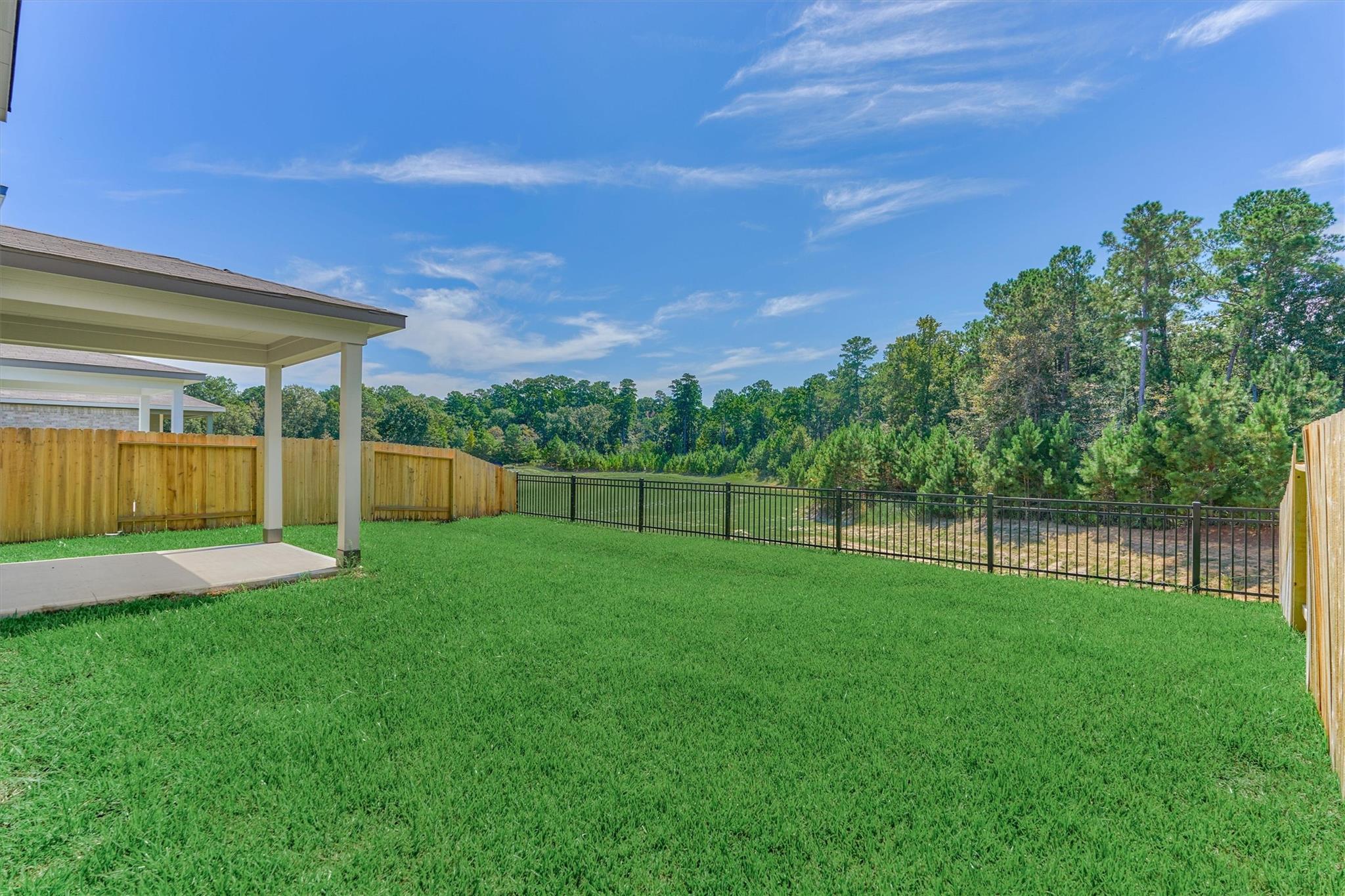 Backyard with a covered patio, fenced lawn, and trees beyond the black metal fence.