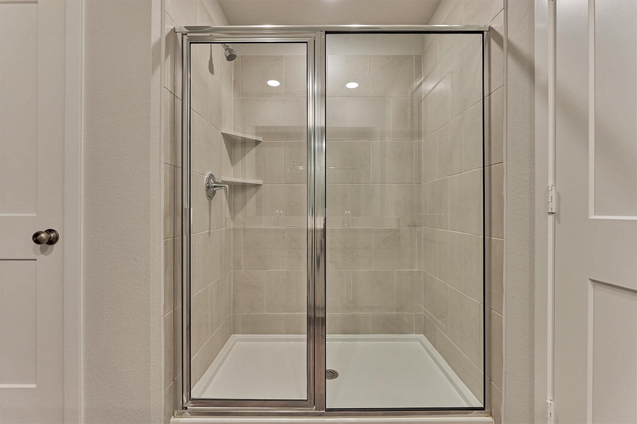 Walk-in shower with glass doors, beige tile, built-in shelves, and chrome fixtures.