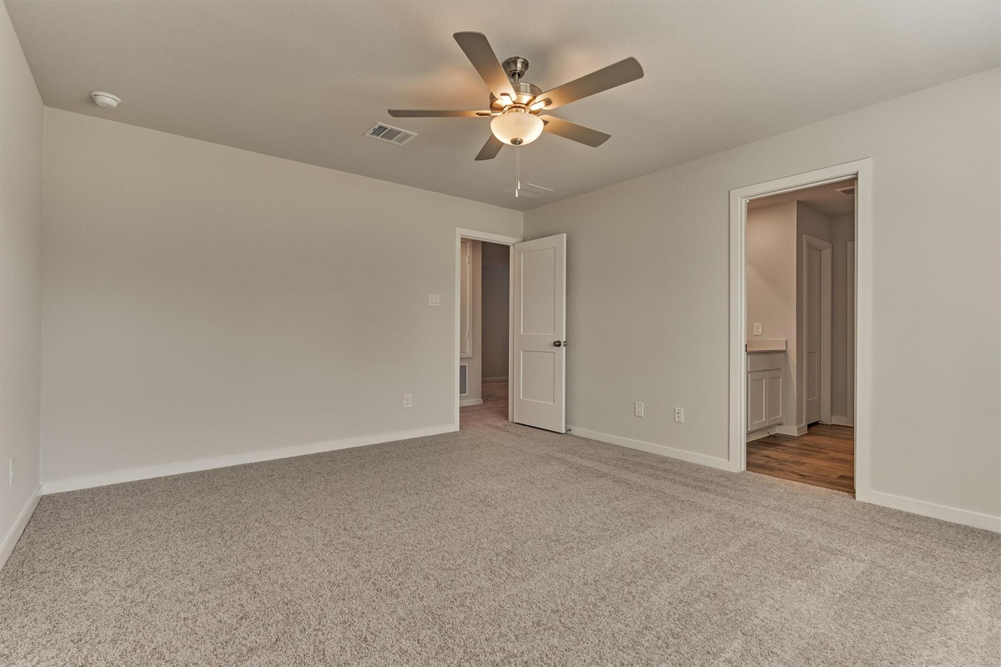 Empty carpeted bedroom with a ceiling fan and open doorways to the hall and bathroom.