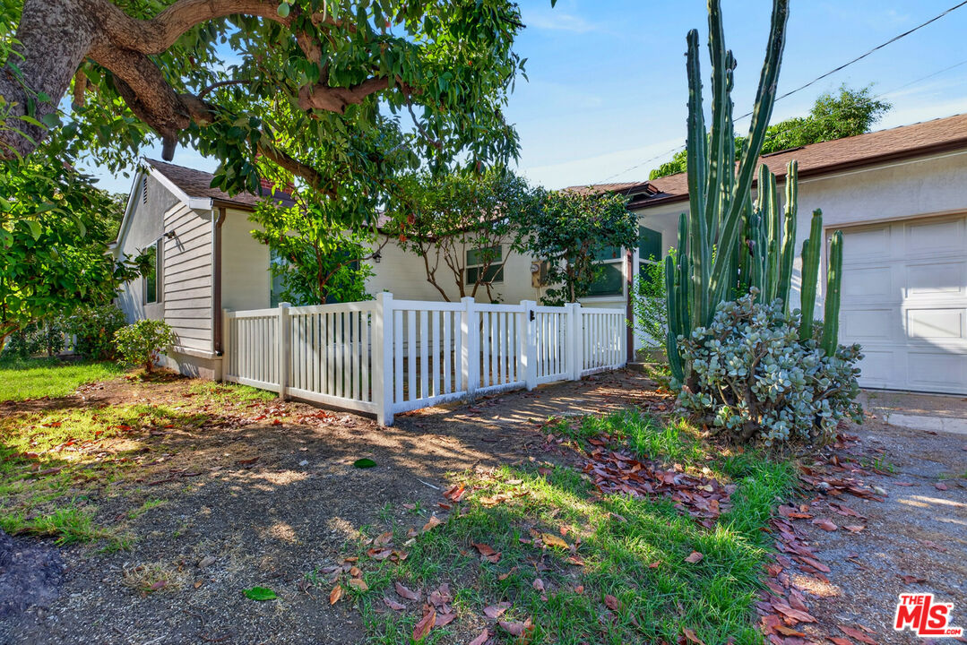 Single-story house behind a white picket fence, framed by leafy trees and tall cacti.
