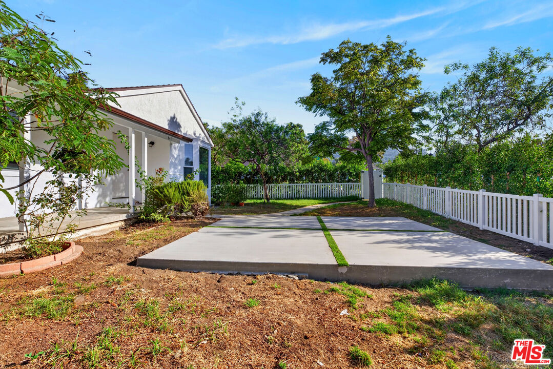 White house beside a broad concrete patio, with a white picket fence and mature trees.