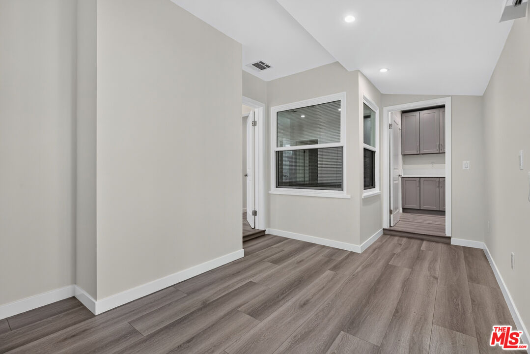 Empty room with gray plank flooring, a window with blinds, and an adjoining area with cabinets.