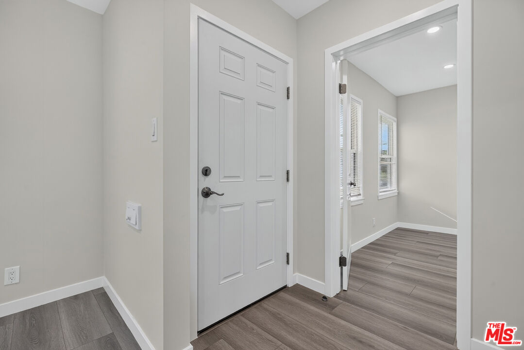 Entryway with a white paneled door and gray plank floors, opening to a bright room with shuttered windows.