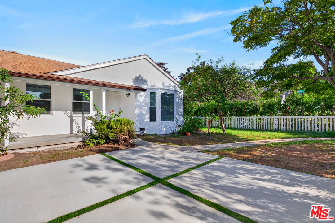 White single-story home with a grass-divided concrete driveway, mature trees, and a white picket fence.