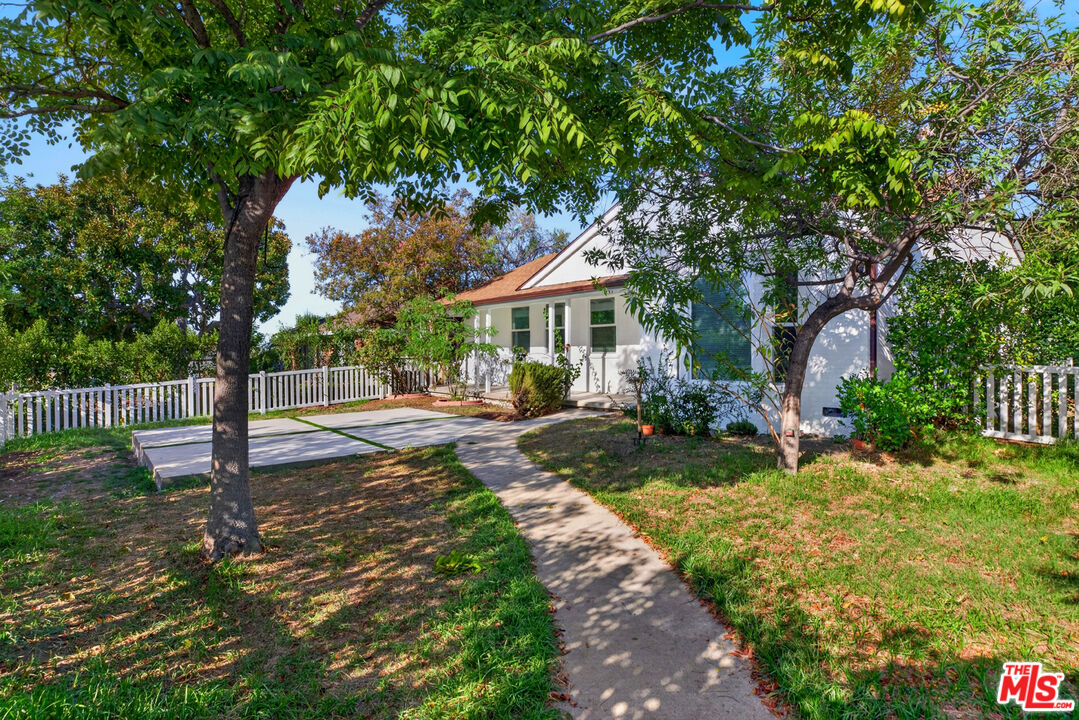 White single-story house with a reddish roof, framed by mature trees and a white picket fence.