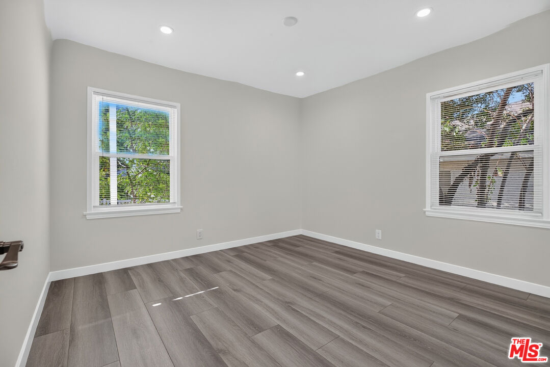 Vacant room with gray plank flooring, recessed lights, and two windows overlooking trees.