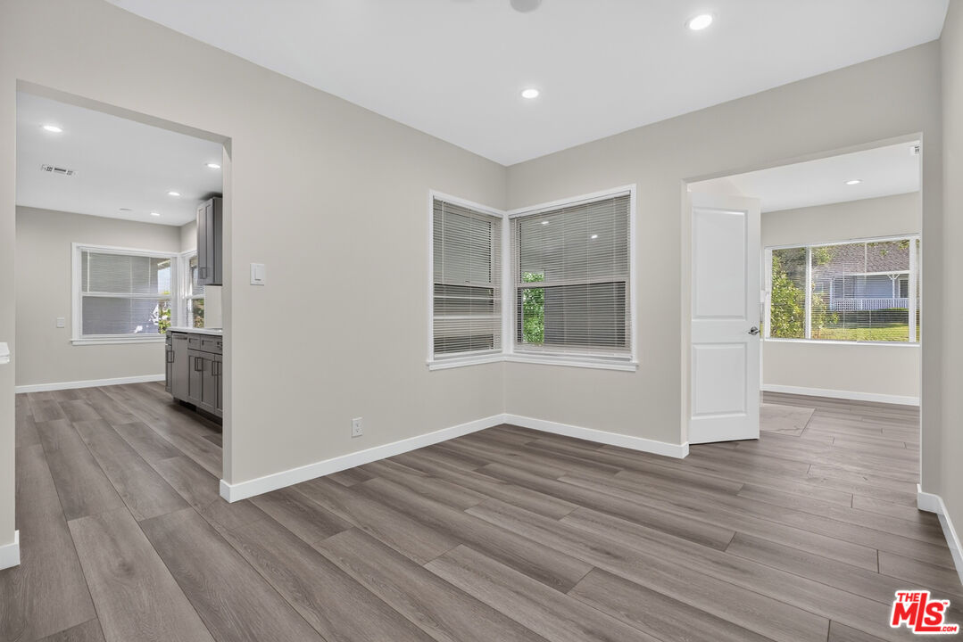 Vacant room with gray plank flooring, corner windows, and openings to the kitchen and adjoining room.
