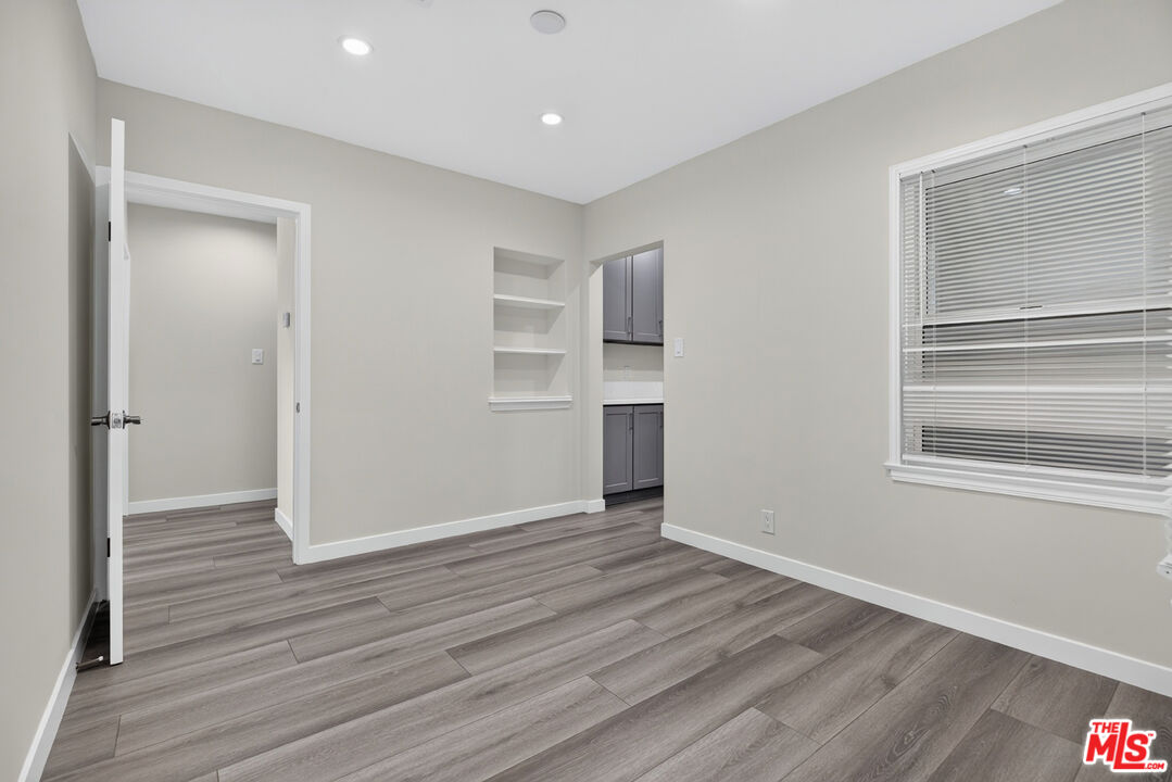Empty room with gray plank flooring, window blinds, built-in shelves, and a glimpse of the kitchen.