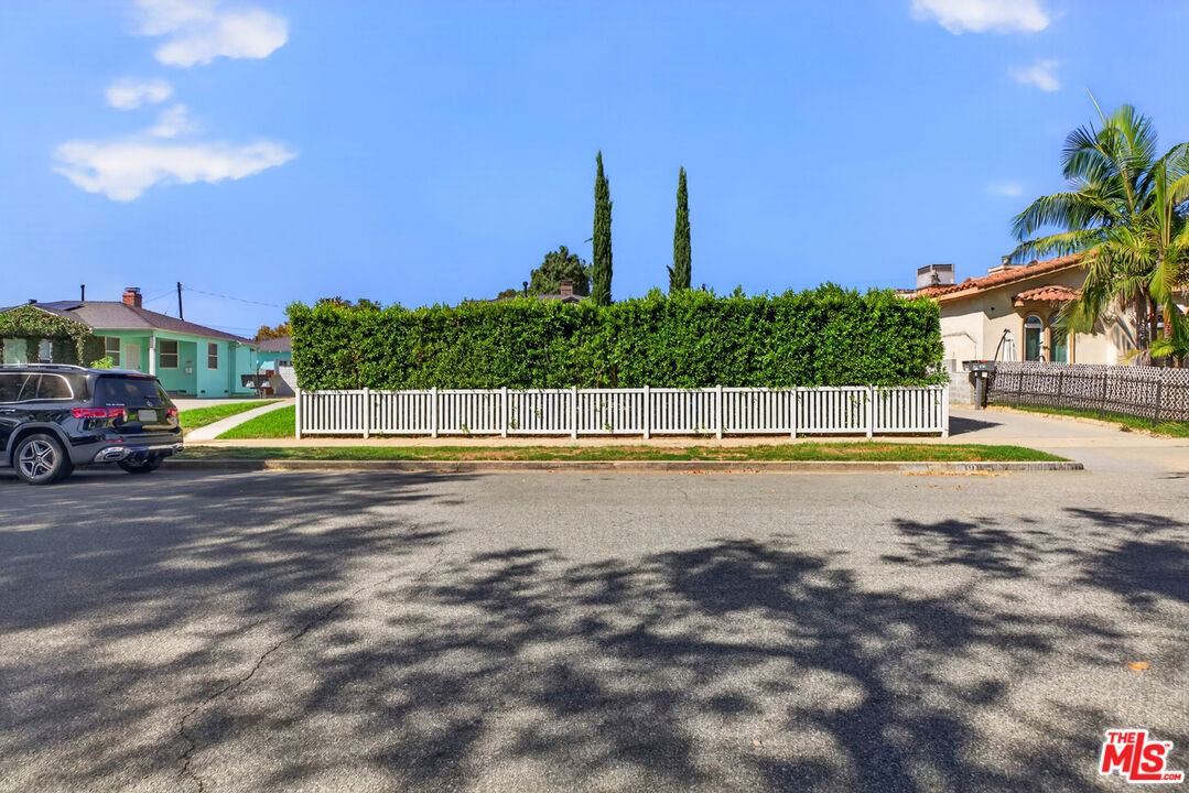 Street-facing home screened by a tall hedge and white picket fence, with palm trees nearby.