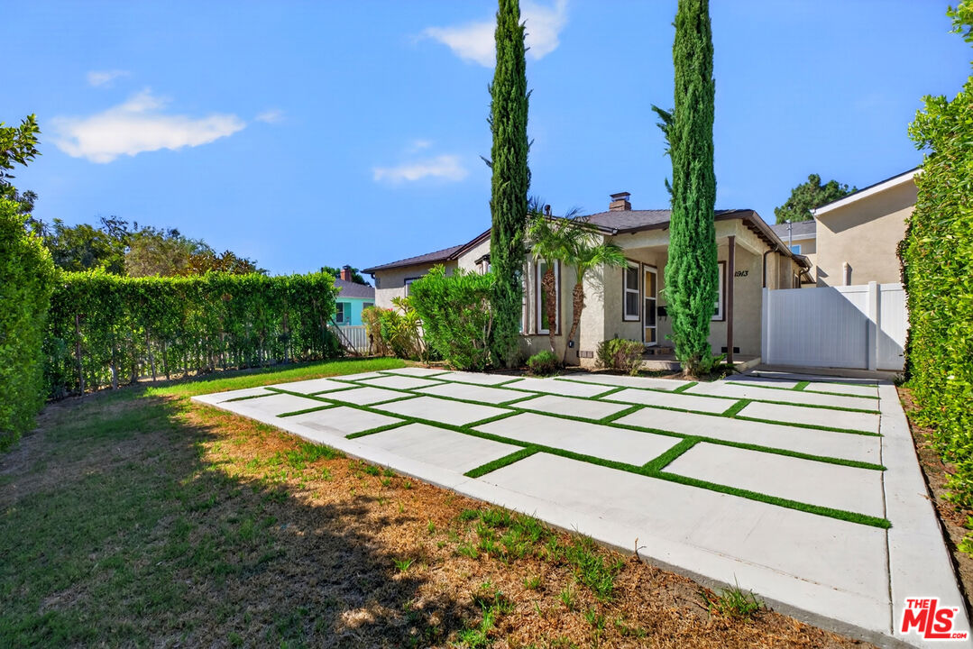 Single-story stucco home with a grass-jointed concrete driveway, tall cypress trees, and a white side gate.