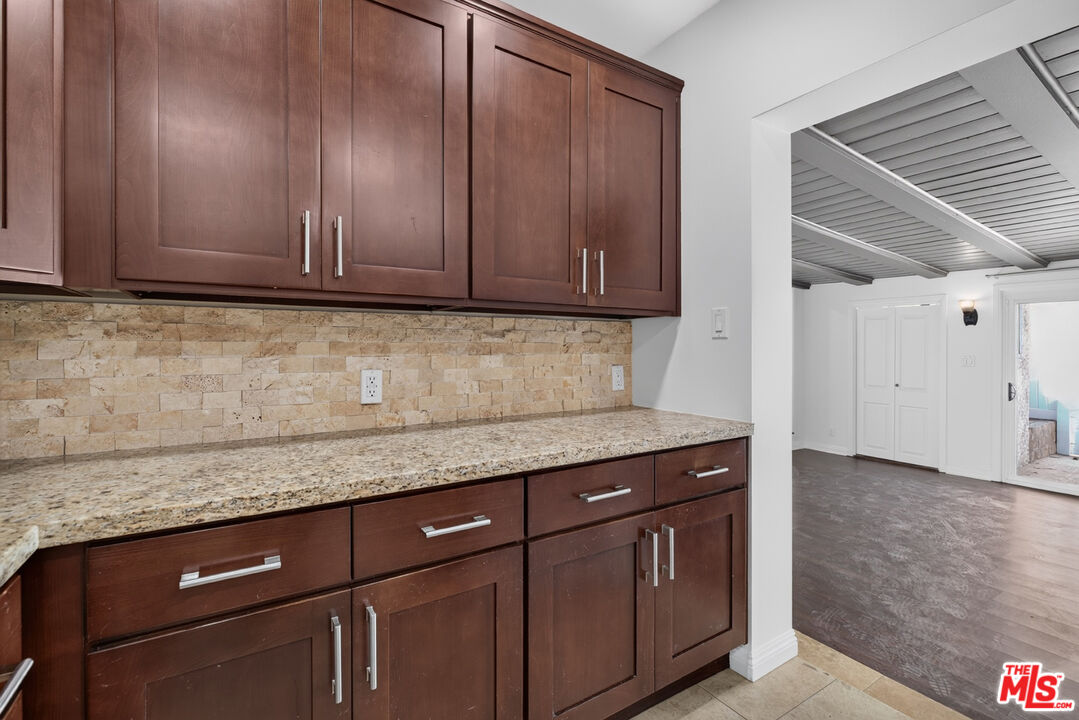 Kitchen with dark wood cabinets, granite counters, and a stone-tile backsplash beside an open adjoining room.