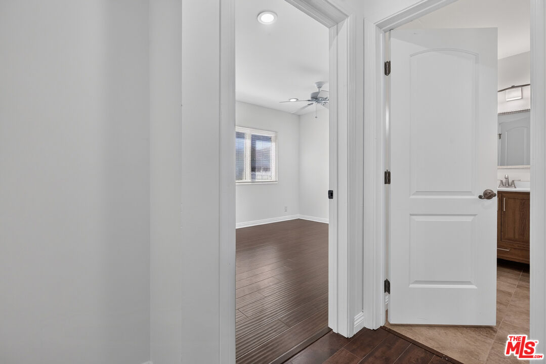 Empty bedroom with dark wood-look flooring and ceiling fan, viewed beside an open bathroom doorway.