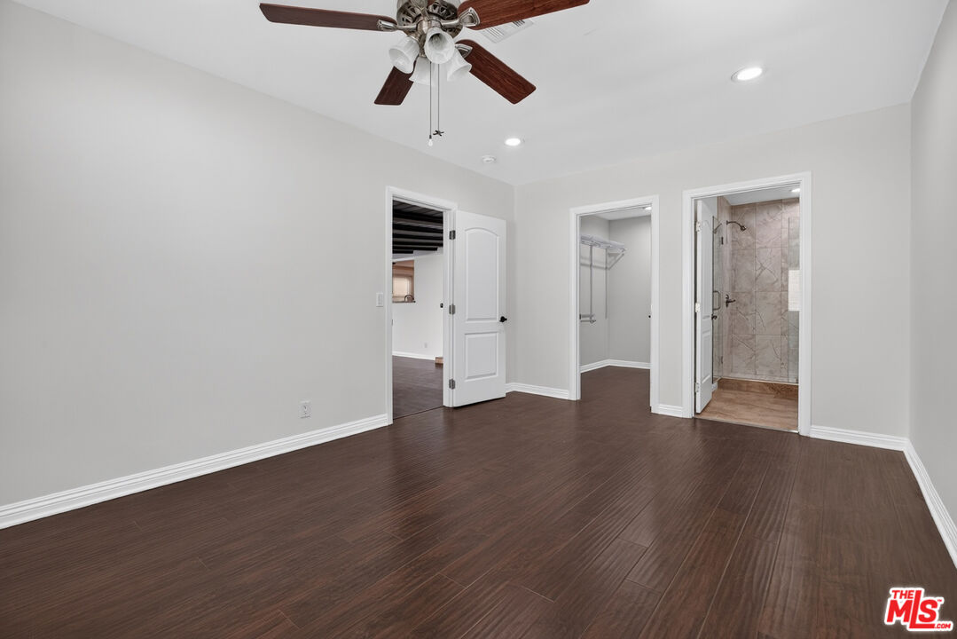 Vacant bedroom with dark wood floors, a ceiling fan, and open doorways to a closet and tiled shower.