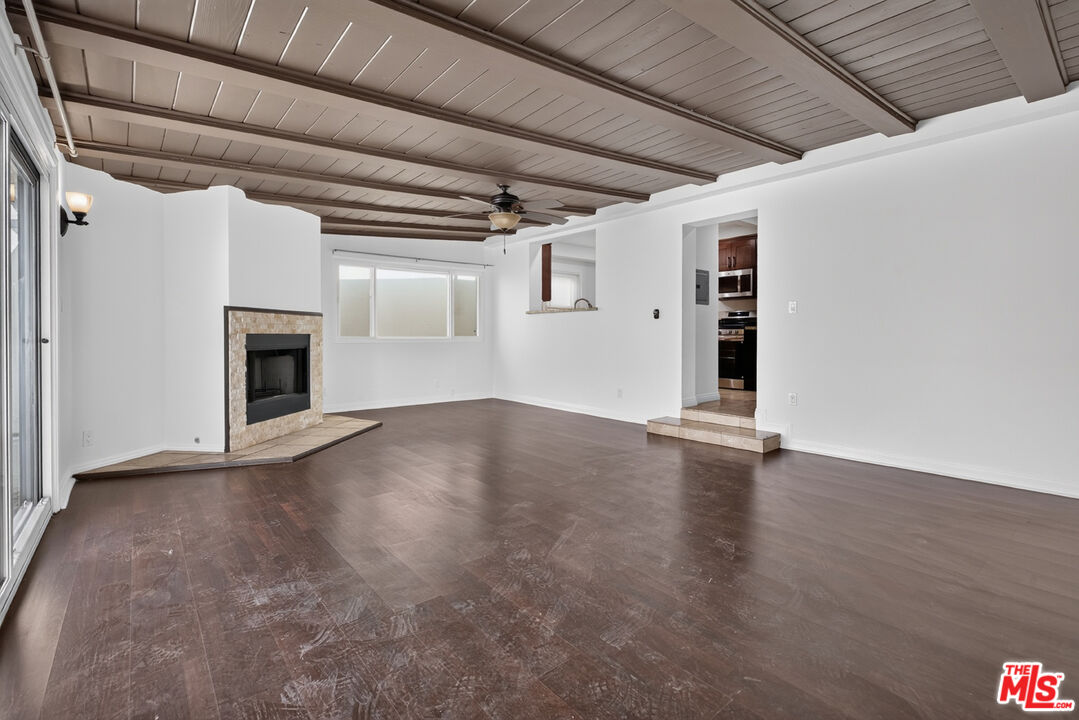 Empty living room with wood-beam ceiling, stone fireplace, and dark wood flooring.