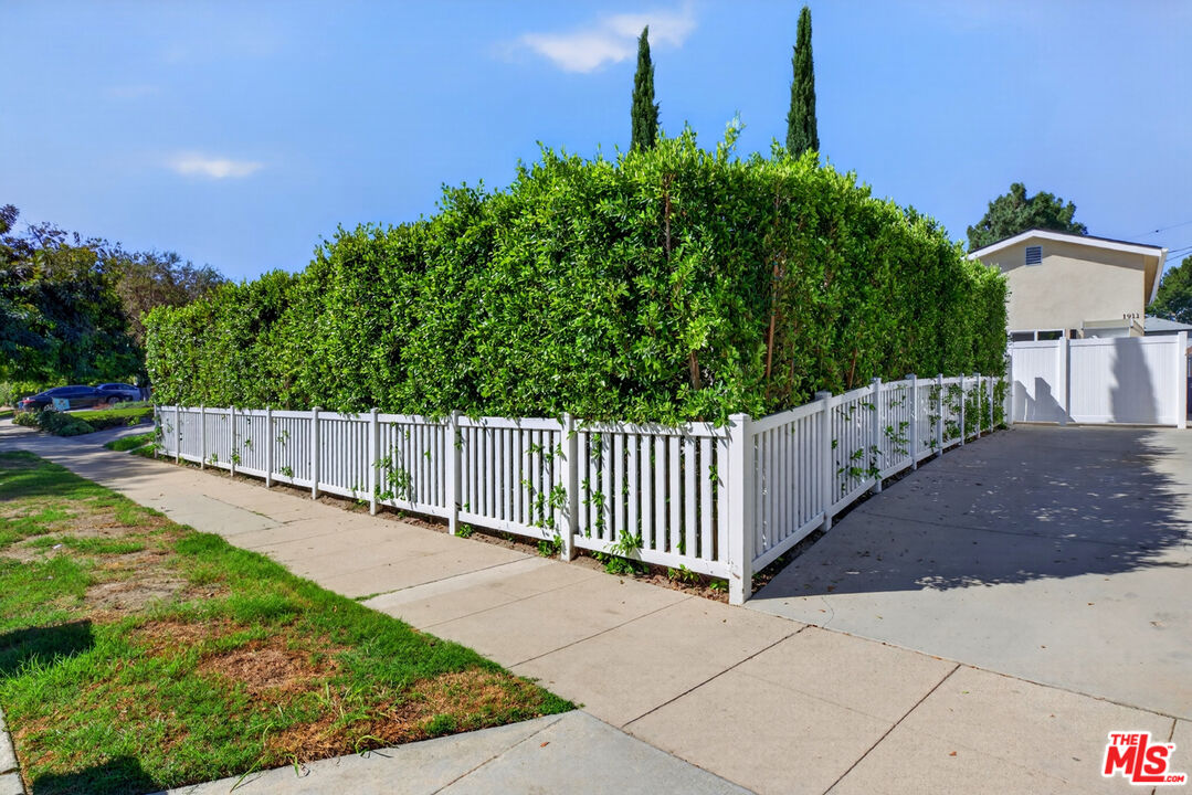 Property exterior with a tall hedge behind a white picket fence and a concrete driveway.