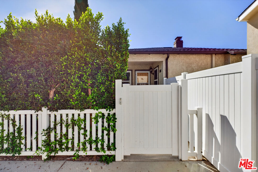 Home entrance behind a tall white gate, bordered by a leafy hedge.