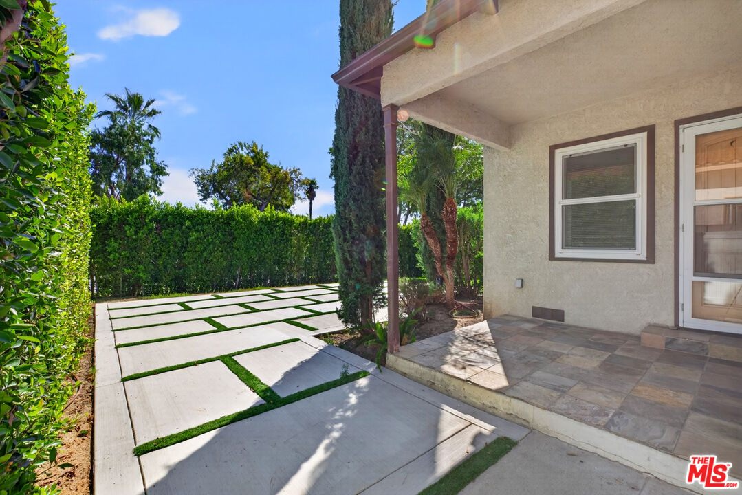 Covered tiled porch beside a geometric concrete walkway with grass strips and landscaped hedges.