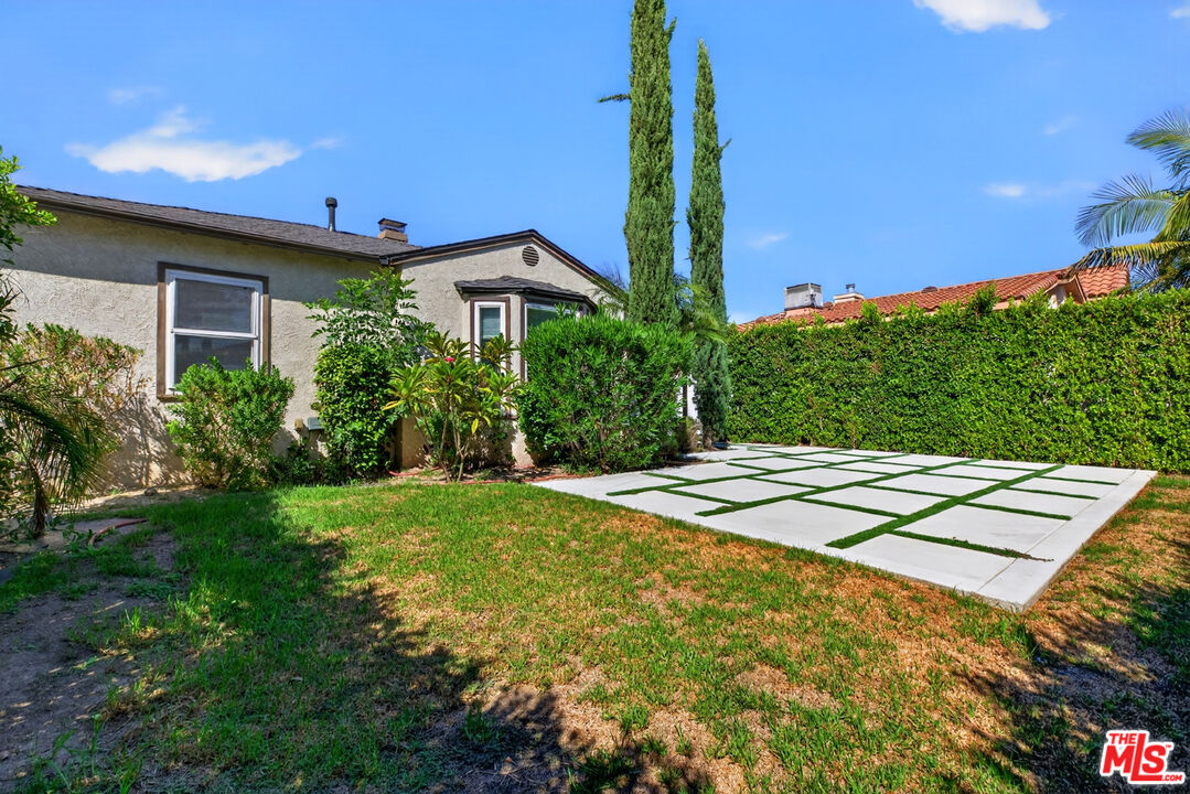 Single-story stucco house beside a grass-jointed concrete parking pad and hedge-enclosed yard.