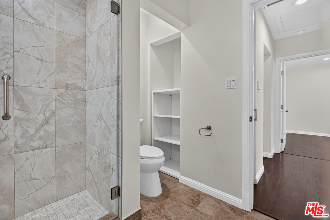 Bathroom with a marble-pattern tiled shower, toilet, and built-in shelving beside a dark-floored hallway.