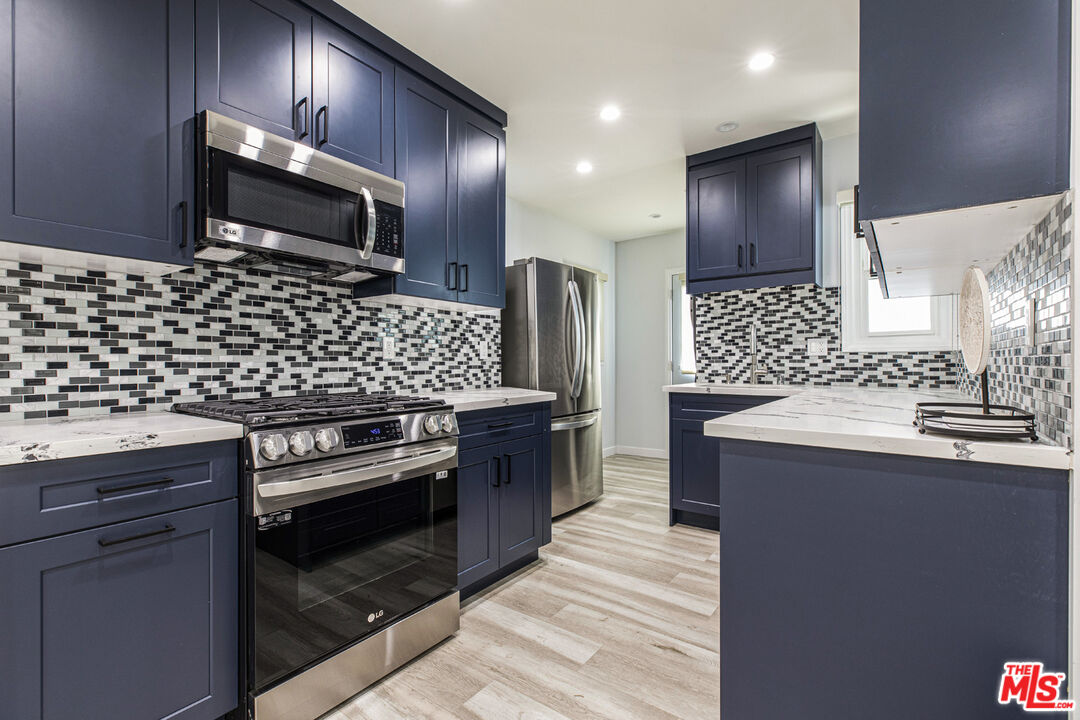 Kitchen with navy cabinets, stainless steel appliances, mosaic backsplash, and light wood-look flooring.