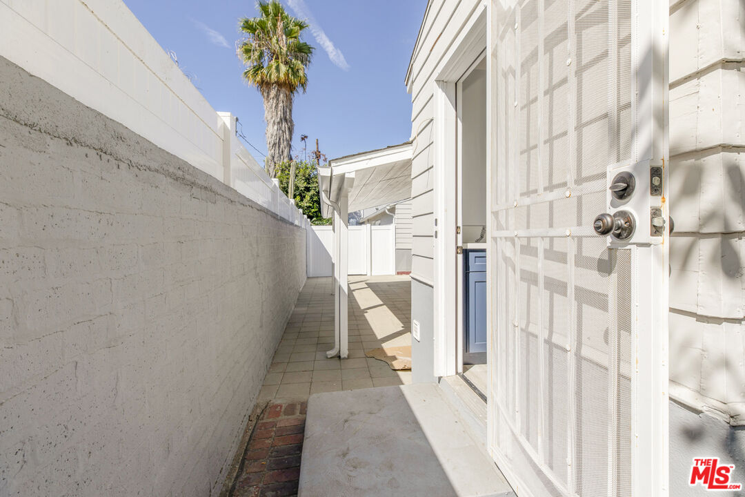 Side entrance walkway beside a white house, with a mesh security door and tall concrete wall.