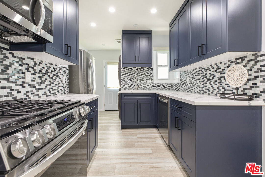 Kitchen with navy cabinets, mosaic tile backsplash, stainless steel appliances, and light stone countertops.