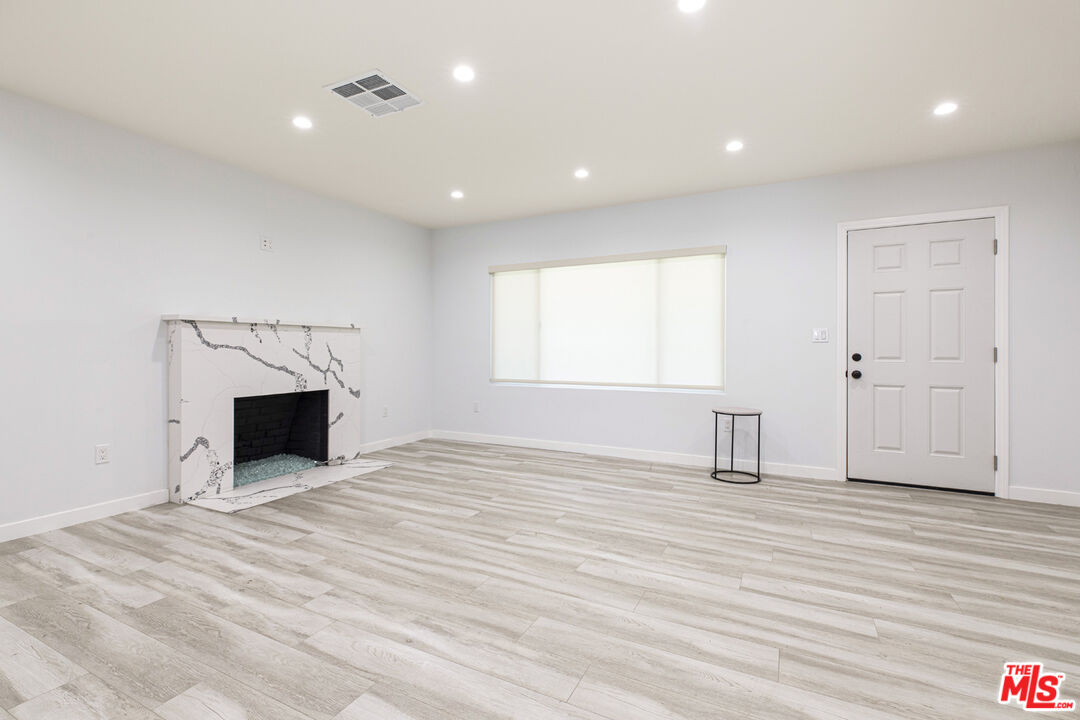 Vacant living room with a marble-pattern fireplace, pale plank flooring, and a wide shaded window.