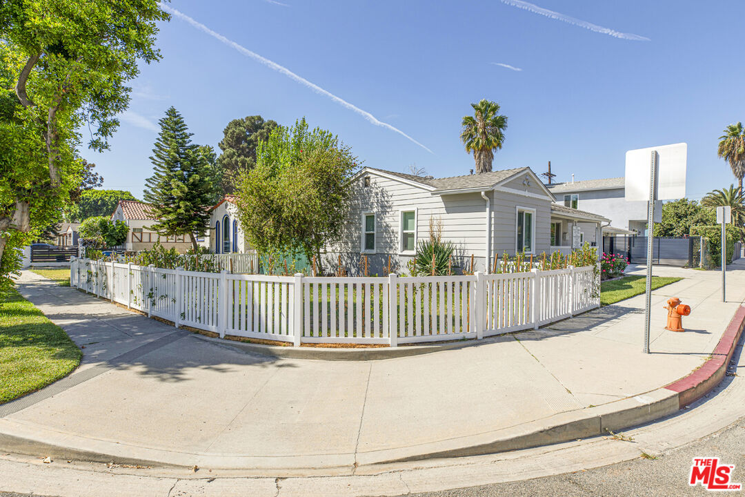 Single-story light-colored house behind a white picket fence at a landscaped street corner.