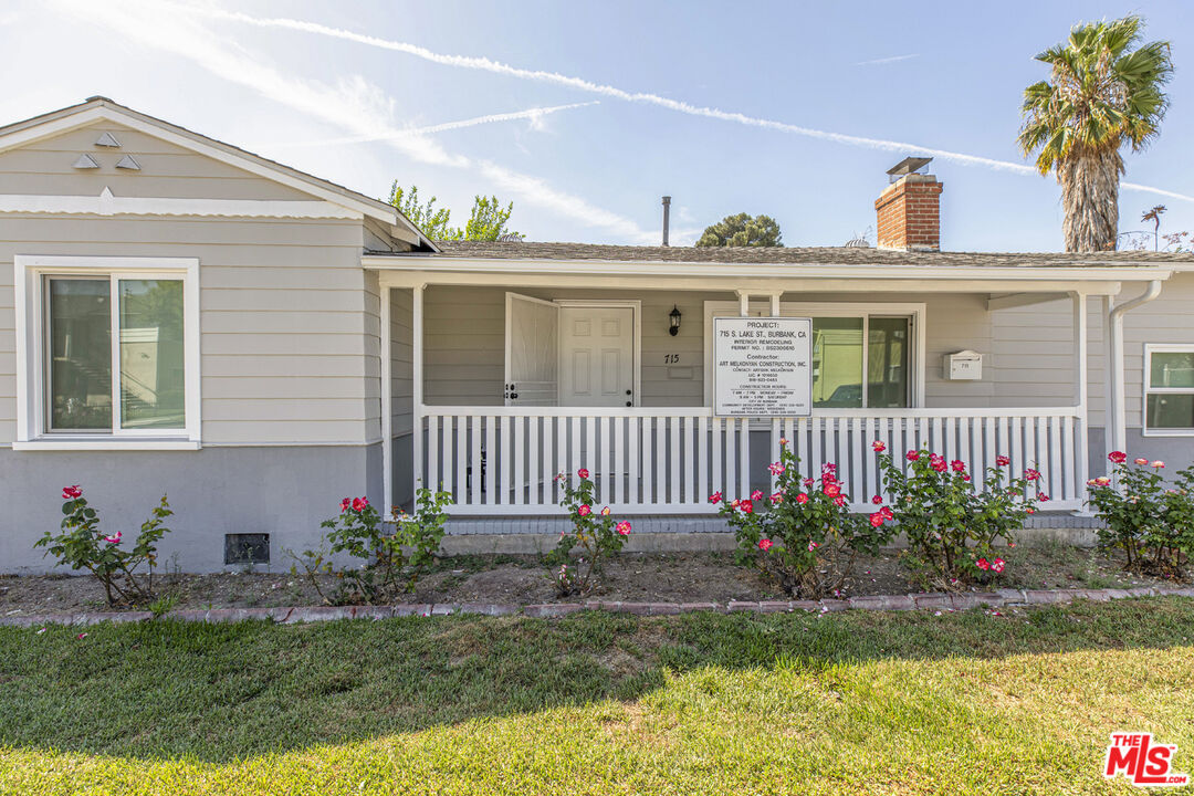 Gray single-story house with a white-railed porch, rose bushes, and a project sign by the entrance.