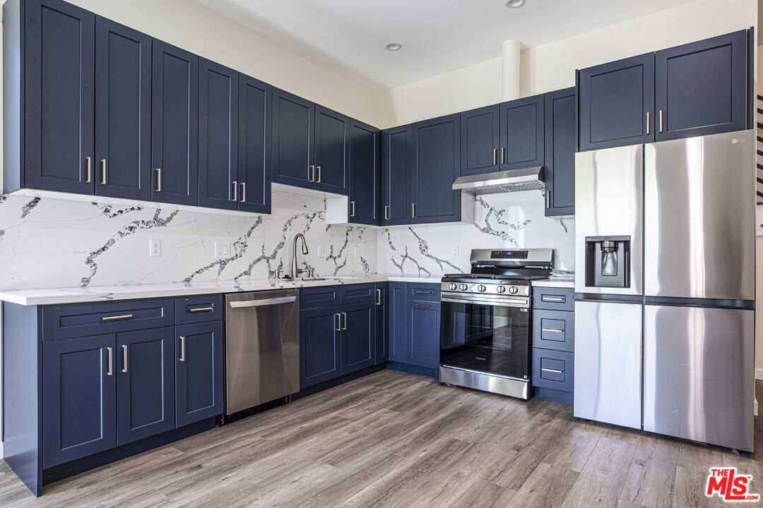 Kitchen with navy cabinets, veined white backsplash, stainless steel appliances, and wood-look flooring.