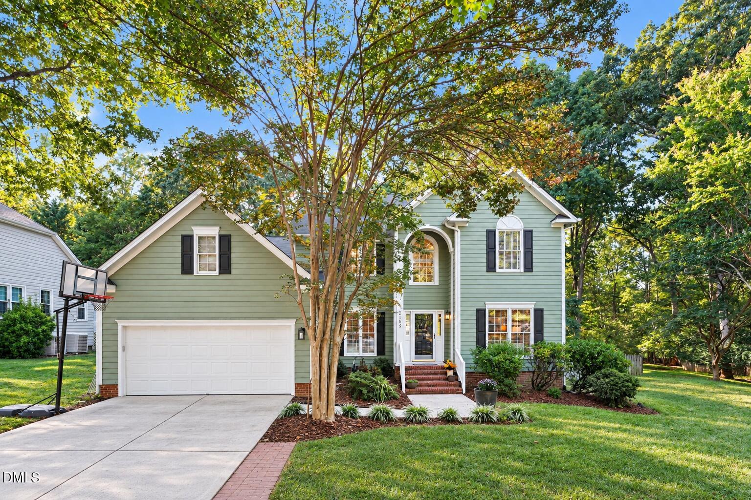 Sage-green two-story house with an attached garage, basketball hoop, and mature trees.